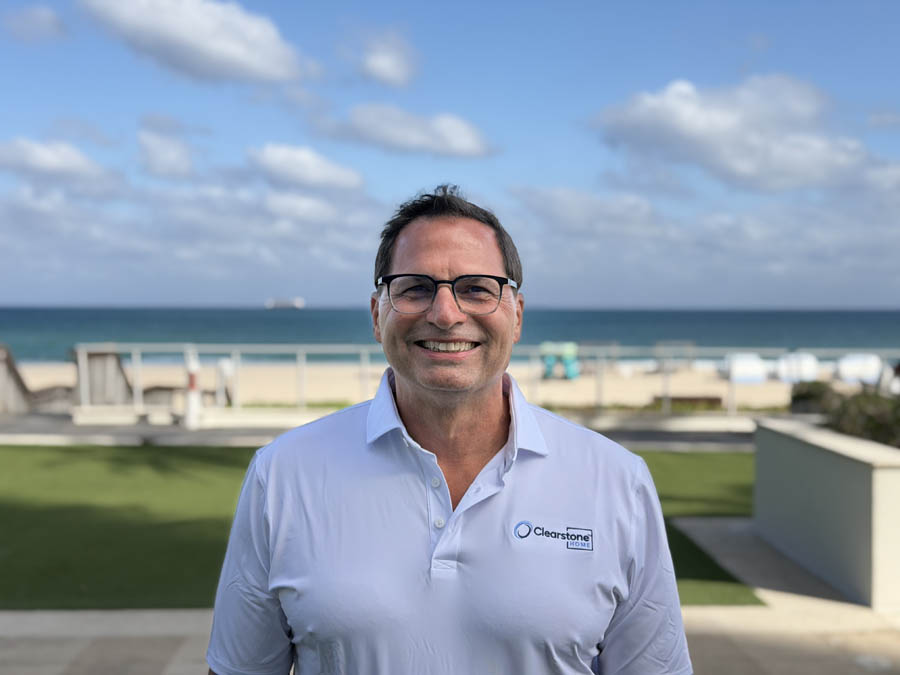 A man in a white shirt with a beach and ocean backdrop under a blue sky.