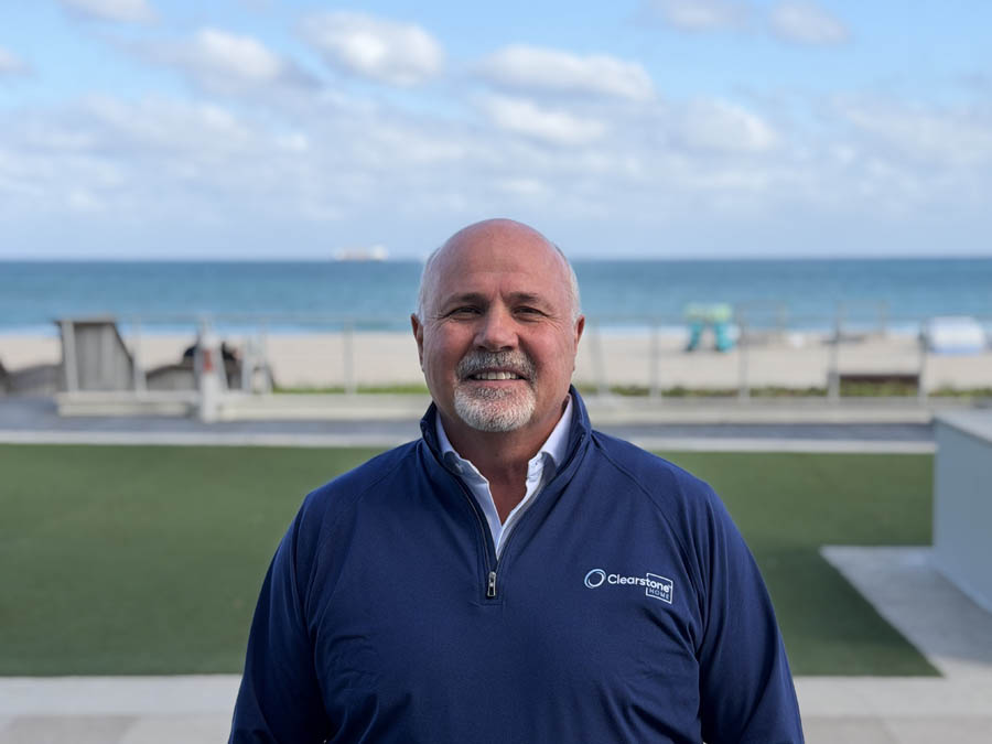 Man in a blue jacket smiling on a beachside patio with a clear sky and ocean in the background.