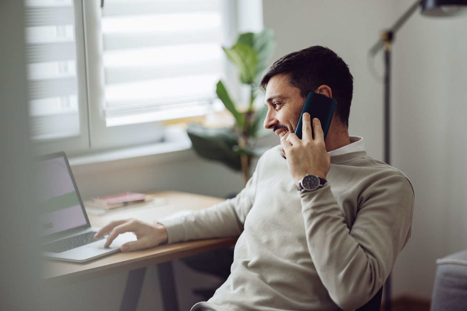 Man on phone working at a home office desk with a laptop, near a window with natural light.